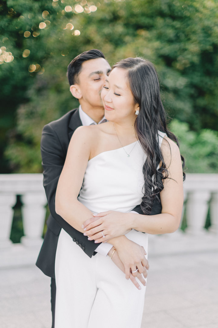 man holds his fiance from behind and plants a kiss on her cheek at their outdoor engagement photography session in Montreal