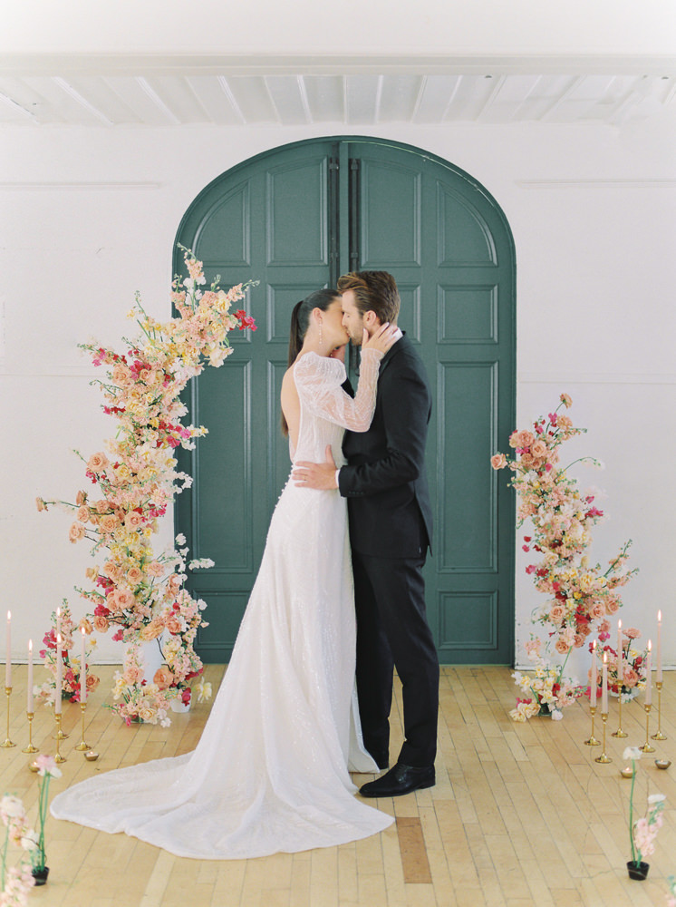 Bride and groom kiss in front of double doors and a floral arch at their wedding