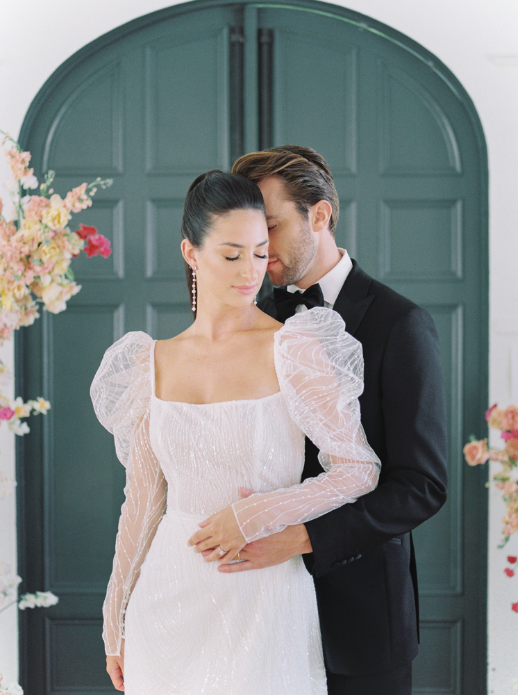 Groom holds his bride in front of a floral arch as they close their eyes for an intimate wedding moment