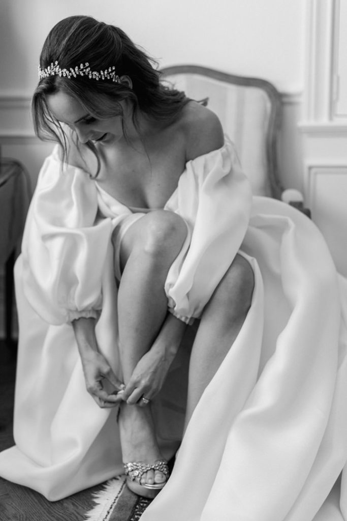 Monochrome photo of a bride in her dress clasping her shoe as she gets ready for her wedding