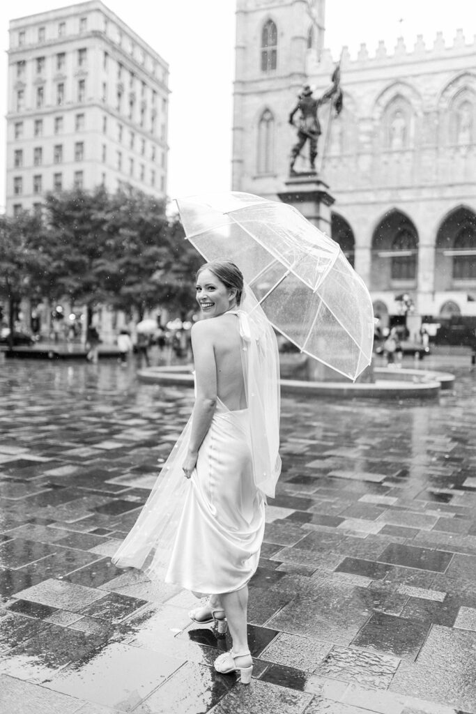 Smiling bride holds up a clear umbrella as captured in a black and white photo by her professional wedding photographer